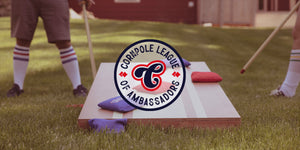 Close up of two people playing cornhole outside on the grass with The Origrinal Cornpole. The Cornpole League of Ambassadors badge overlays the photo.