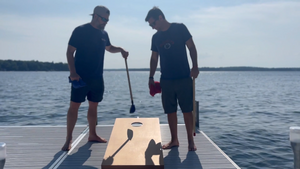 Two men outside on a dock playing cornhole. Ocean can be seen in the background. One man is using The Original Cornpole to pick up cornhole bags.